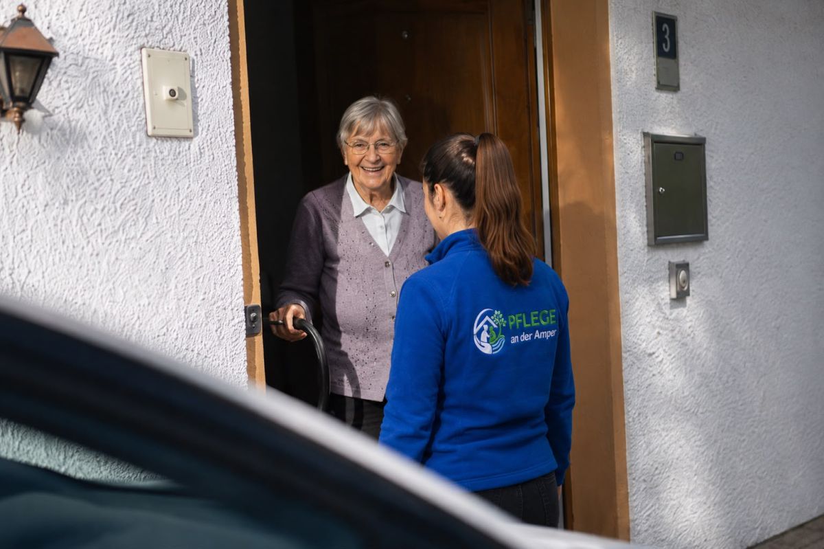 Eine Pflegekraft von Pflege an der Amper wird an der Haustür von einer lächelnden älteren Dame begrüßt.